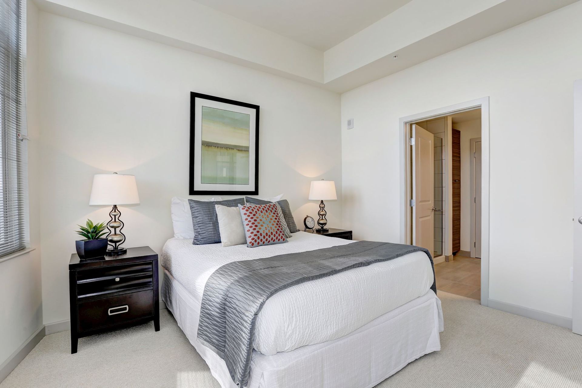 Bright bedroom with a neatly made white bed, gray bedding, bedside lamps, and an open doorway at Solaire 7077 Woodmont in Bethesda, MD.