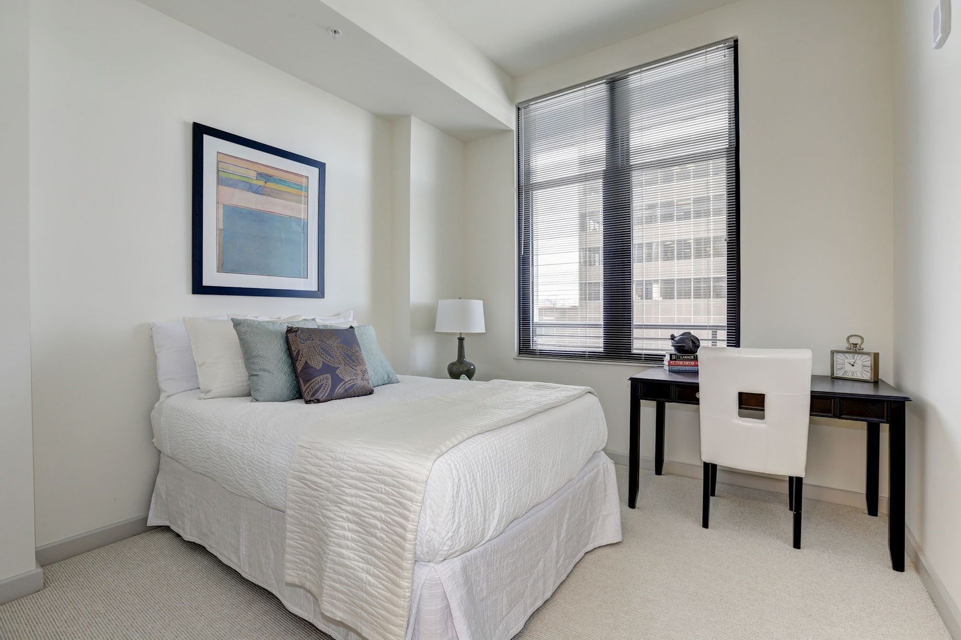 Neat white bedroom with bed, desk, lamp, and window blinds at Solaire 7077 Woodmont in Bethesda, MD.