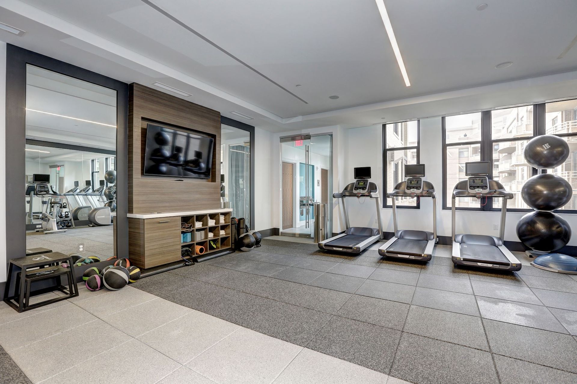 Modern gym with treadmills, dumbbells, wall TV, and mirrored walls in a bright room at Solaire 7077 Woodmont in Bethesda, MD.