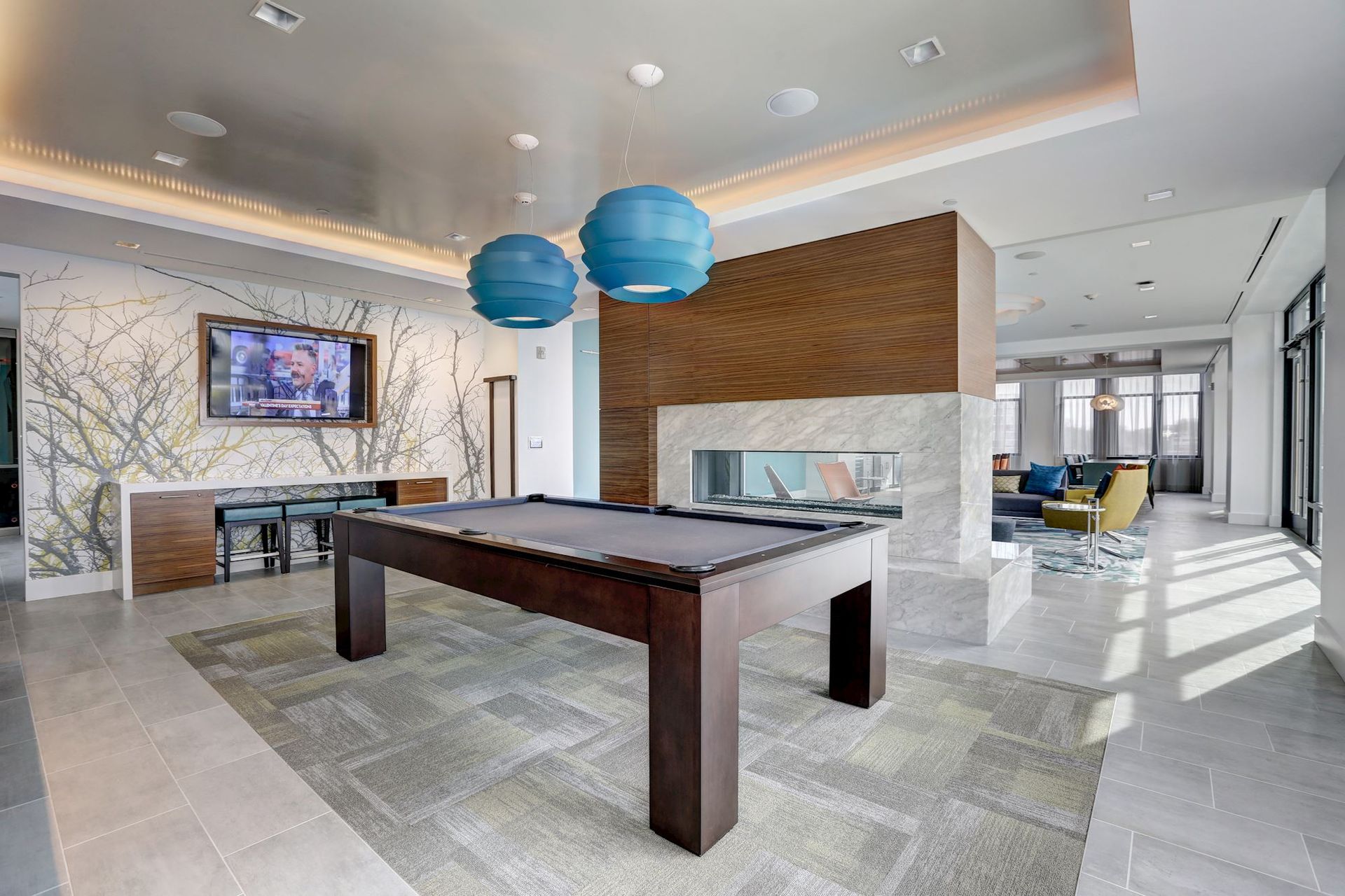 Modern open-plan living room with a pool table, blue pendant lights, fireplace, and TV wall at Solaire 7077 Woodmont in Bethesda, MD.