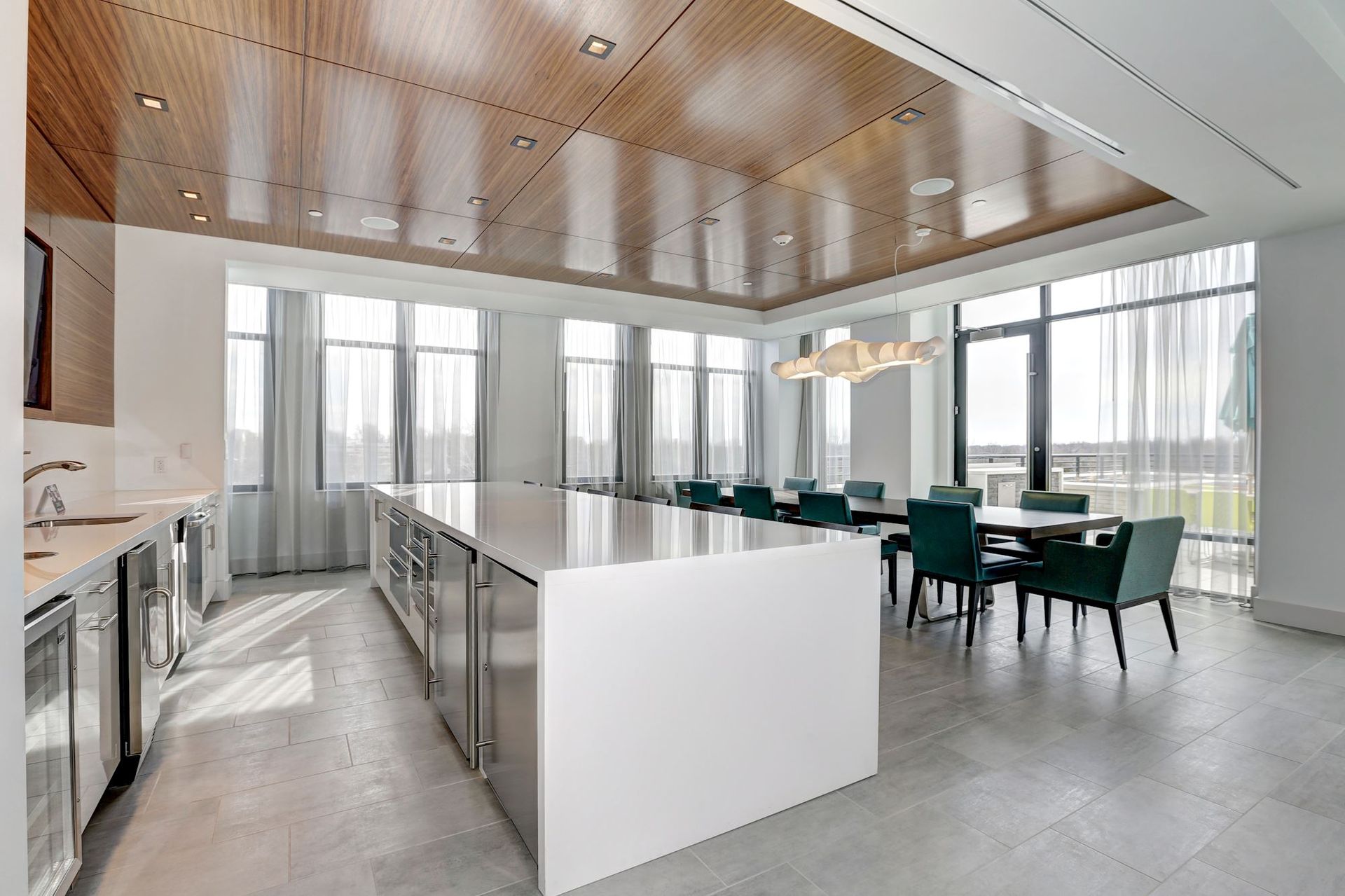 Modern open kitchen and dining area with island, white cabinetry, wood ceiling, and teal chairs by large windows at Solaire 7077 Woodmont in Bethesda, MD.