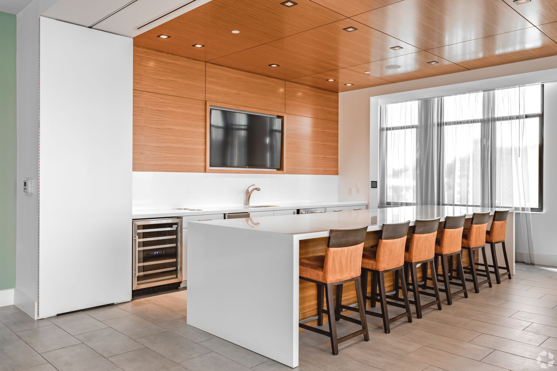 Modern kitchen with white island, wood ceiling, and a row of bar stools by large windows at Solaire 7077 Woodmont in Bethesda, MD.