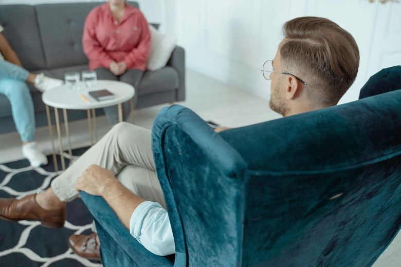 Two people in a living room, one seated on a teal chair facing a person on a gray sofa.