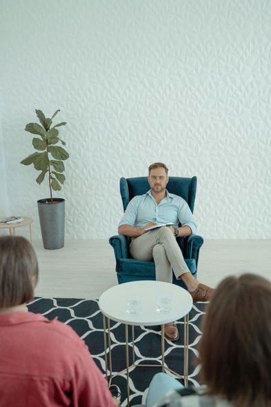 Man sitting in a teal armchair speaking to an audience in a bright room with a plant and round table