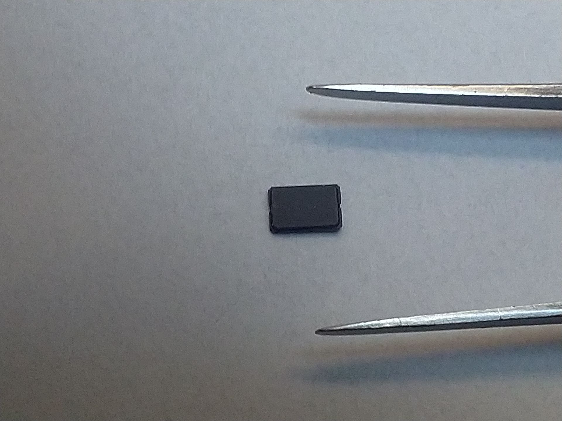 Small black rectangular piece on a gray surface between two metal tweezers.