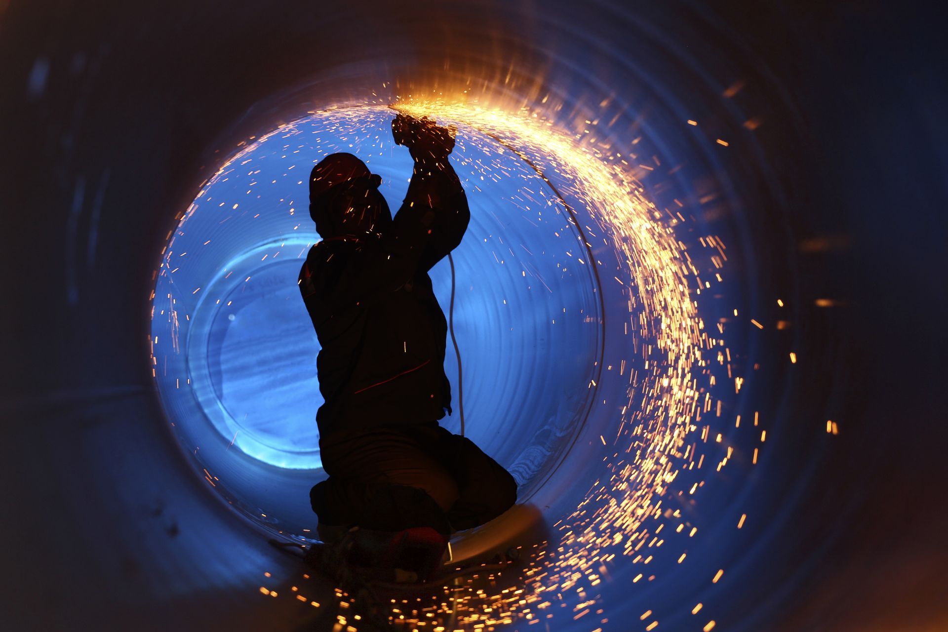 Silhouetted worker sparks inside a glowing blue industrial pipe or tunnel