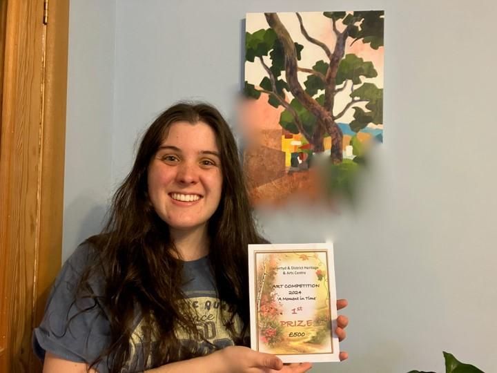 Woman smiling and holding a framed certificate beside a colorful painting on a wall