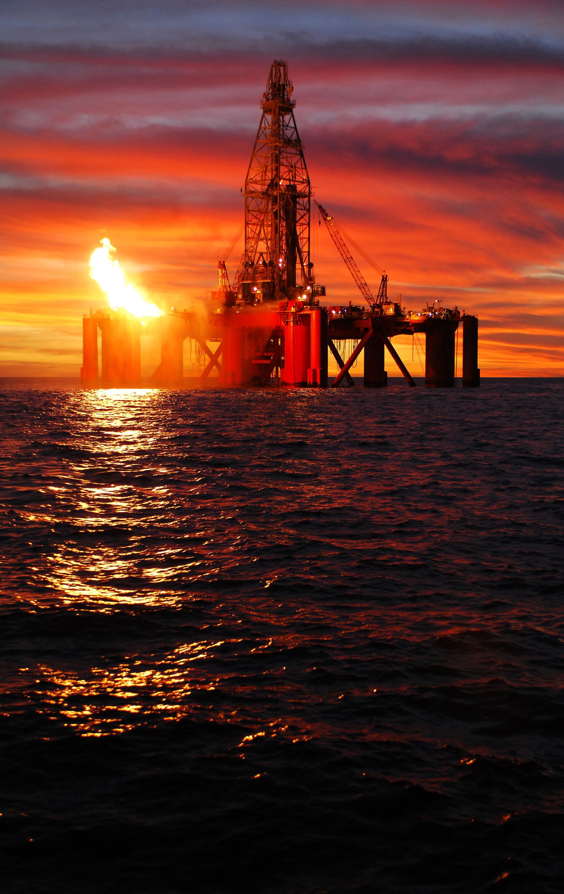 Oil rig silhouetted on the sea at sunset, with fiery orange sky and reflected light on the water