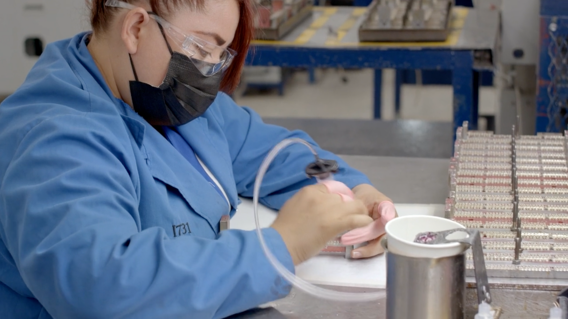 Worker in blue lab coat and mask carefully handling small parts at a workbench in a factory