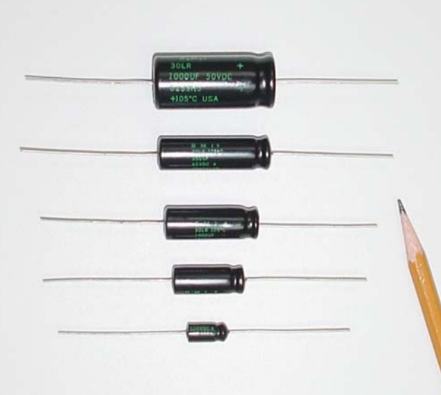 Five black cylindrical capacitors of decreasing size lined up on a white background with a pencil beside them