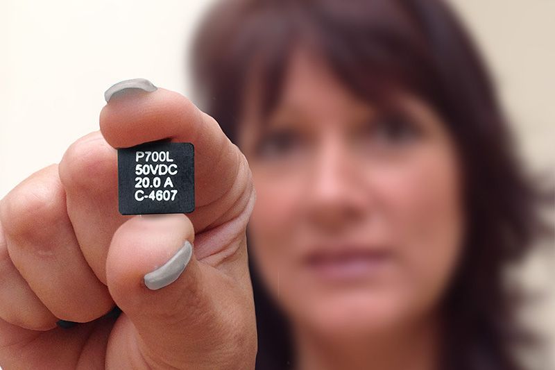 Hand holding a small black electronic component toward the camera, with a blurred person in the background