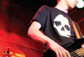 Person playing bass guitar onstage in a black shirt with a white skull graphic, red lighting background