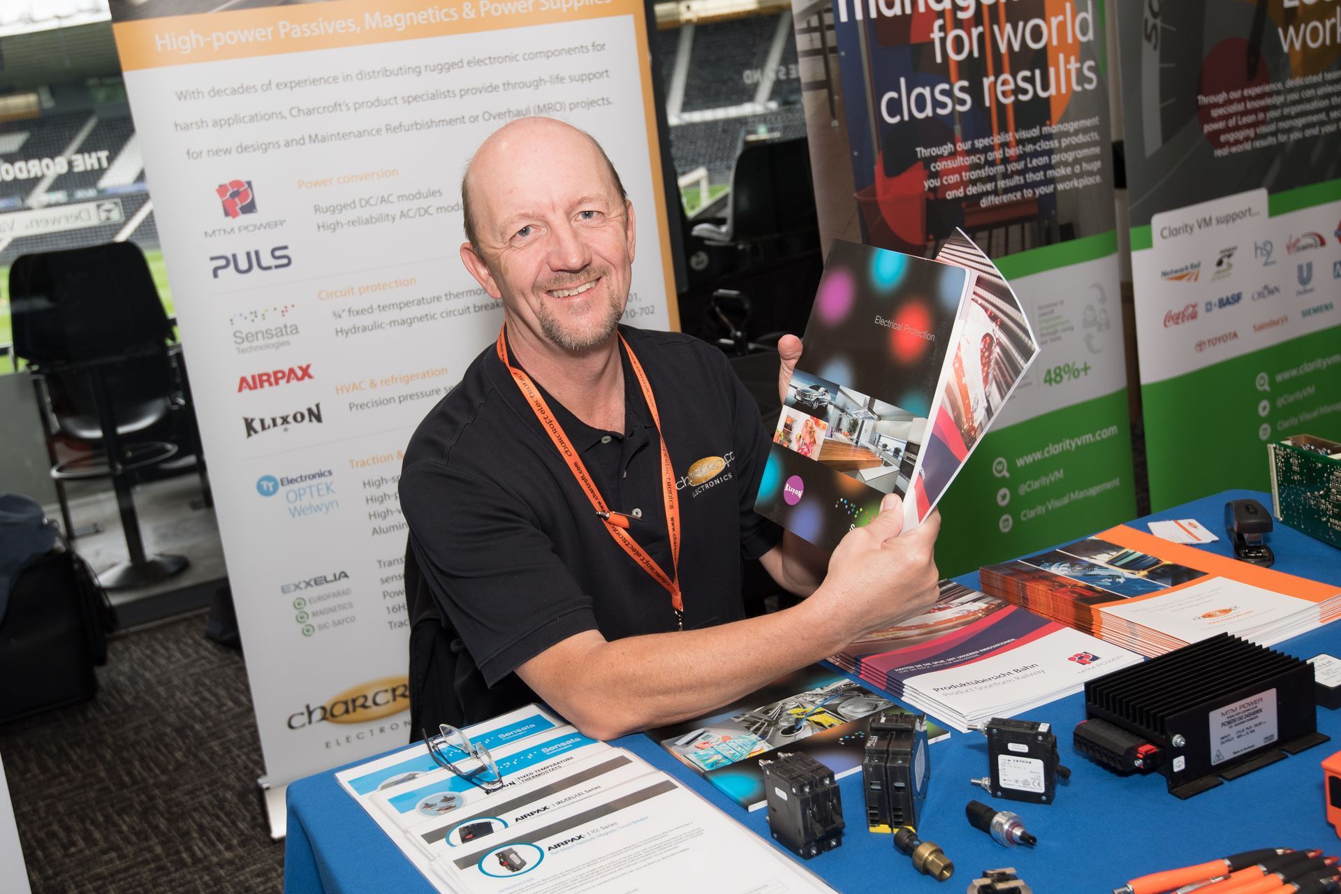 Smiling exhibitor at a trade show booth, holding brochures beside product displays and banners