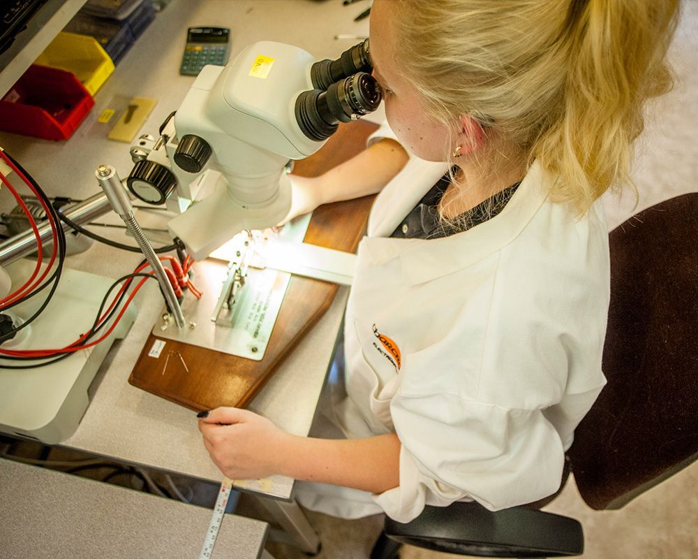 Person in a lab coat using a microscope at a workbench with wires and tools.