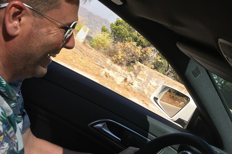 Man driving a car, smiling out the window on a sunny roadside.