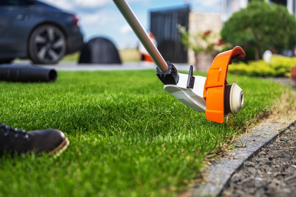 Person edging a lawn with a trimmer; green grass, black edging, orange and white trimmer.