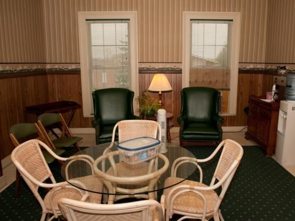 A cozy lounge with wicker chairs around a glass table, two green armchairs, lamp, and windows with blinds.