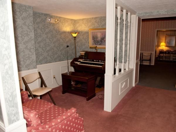 Cozy sitting room with red carpet, floral chair, organ, floor lamp, and white half-wall opening to a bedroom.