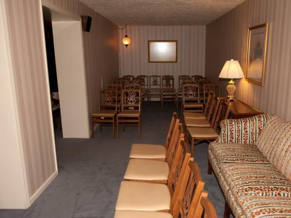 Dim room with rows of wooden chairs, blue carpet, lamps, and patterned sofa along the wall.