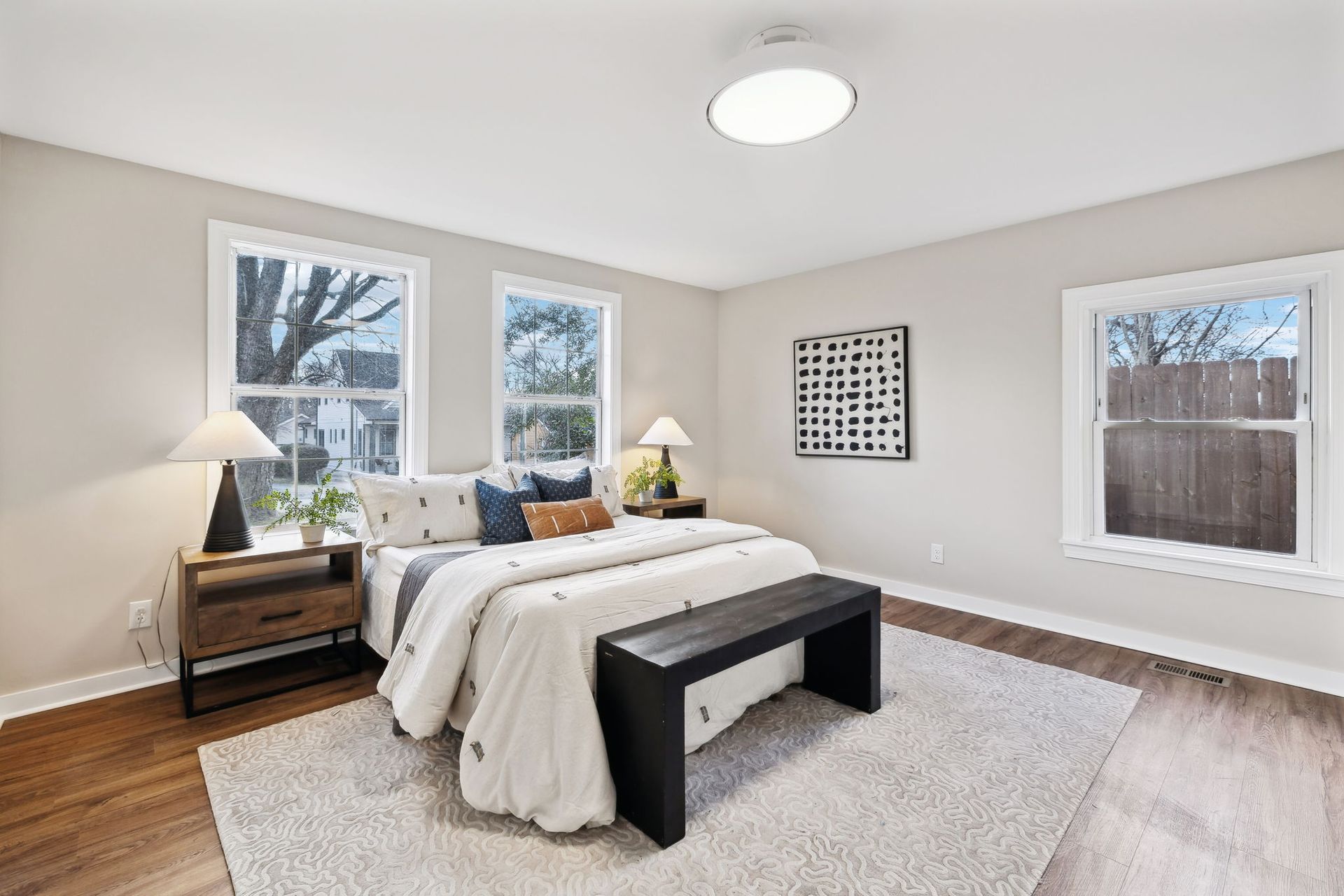 A bedroom with a bed , bench , nightstand , and two windows.