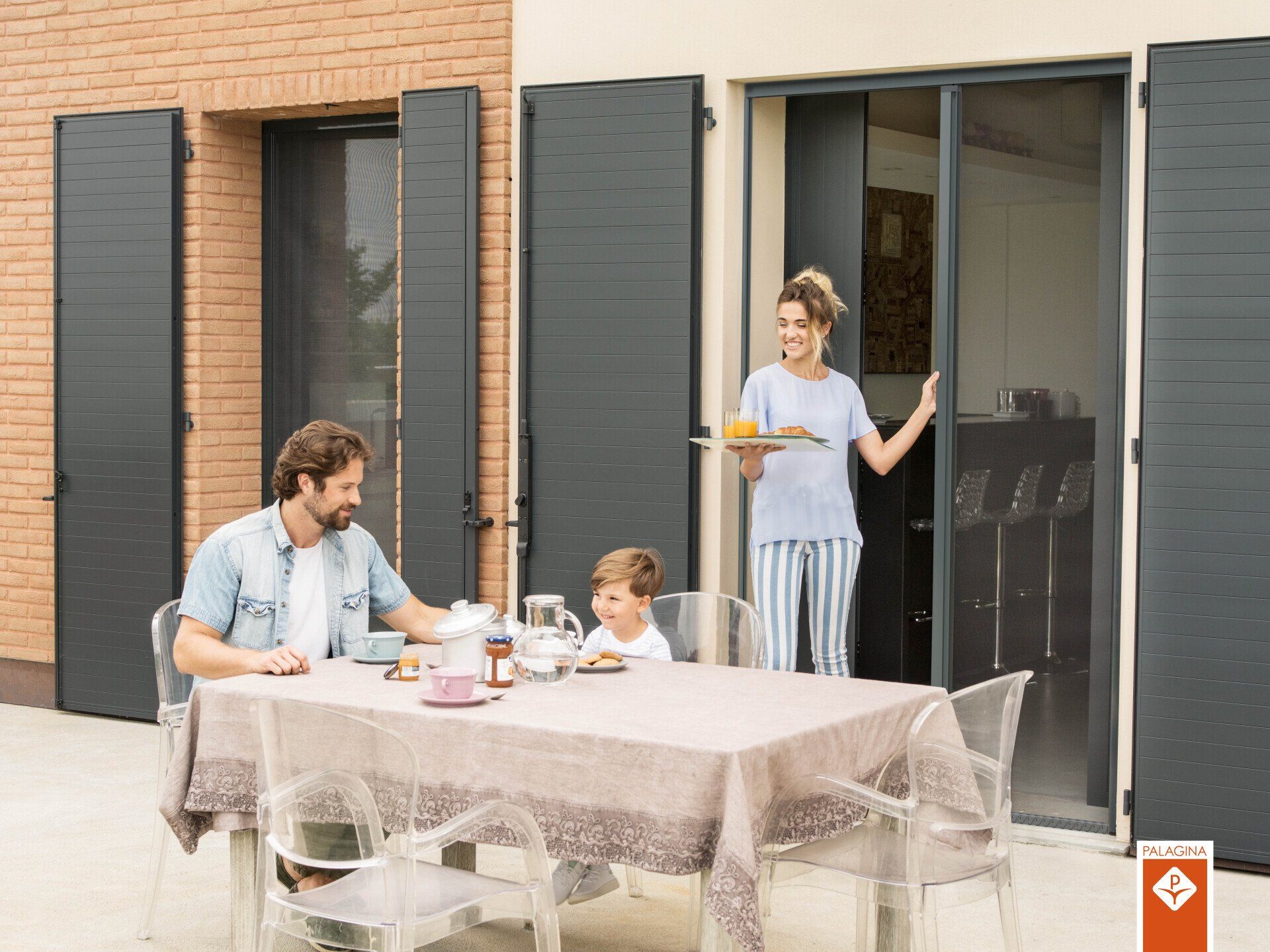 Famiglia che fa colazione in terrazza. Donna con vassoio, porta scorrevole aperta, persiane grigie, sedie trasparenti, sole.
