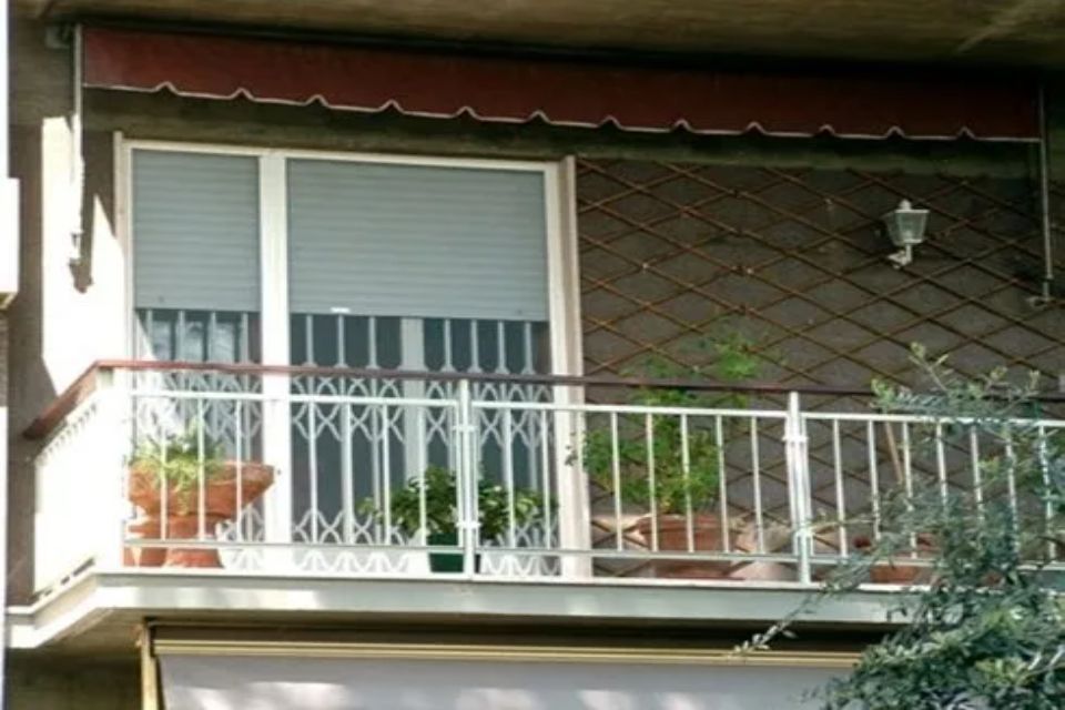 Balcone con ringhiera bianca, piante in vaso, finestra con persiana chiusa e tenda da sole rossa.