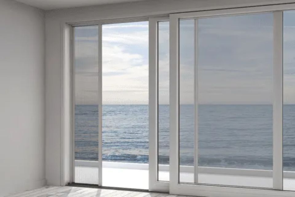 Porte scorrevoli in vetro si aprono sulla vista dell'oceano. Infissi bianchi, cielo coperto.