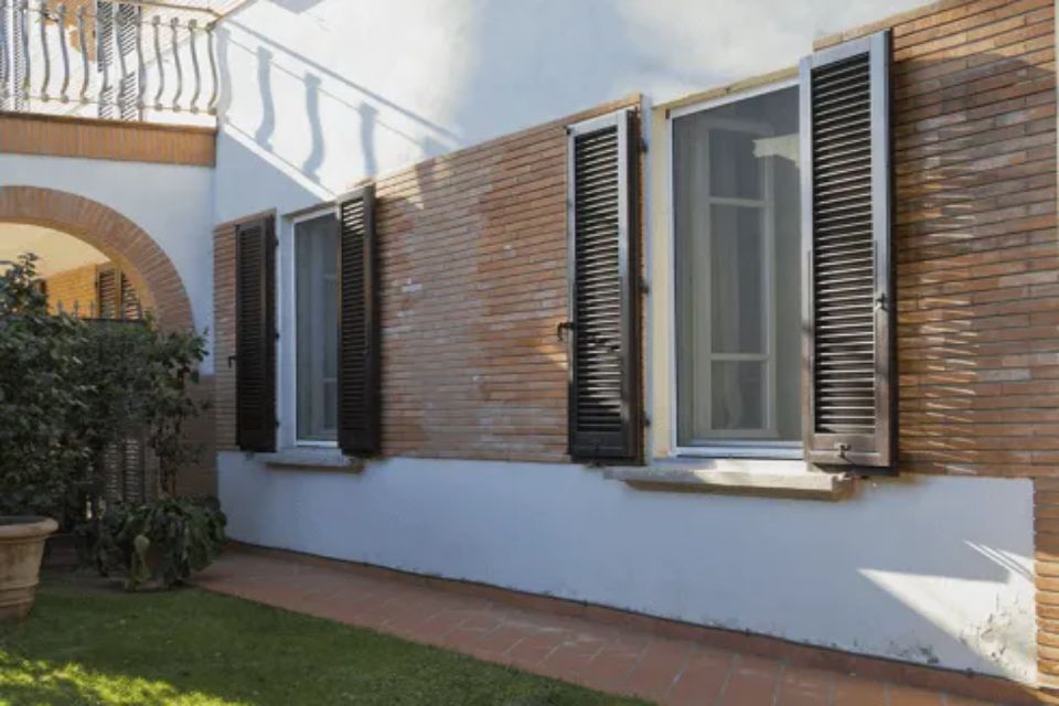 Vista esterna di un edificio con persiane nere aperte alle finestre, muro di mattoni, facciata bianca e vegetazione.