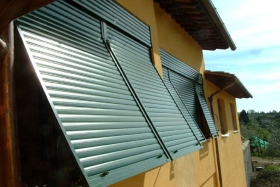 Esterno di un edificio con tre persiane metalliche verdi aperte che proiettano ombre.