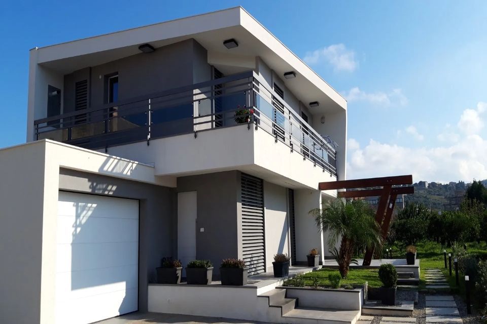 Moderna casa a due piani con esterni bianchi e grigi, balcone, garage e giardino paesaggistico nelle giornate di sole.