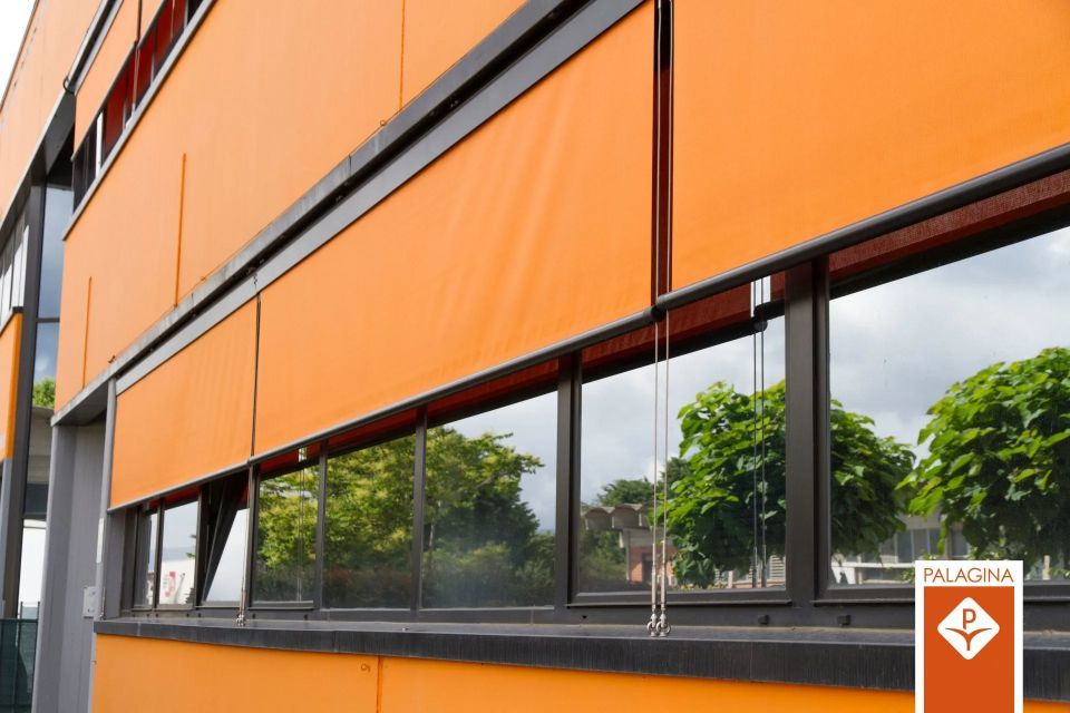 Edificio arancione con parasole arancioni retratti sopra le finestre riflettenti; alberi riflessi.