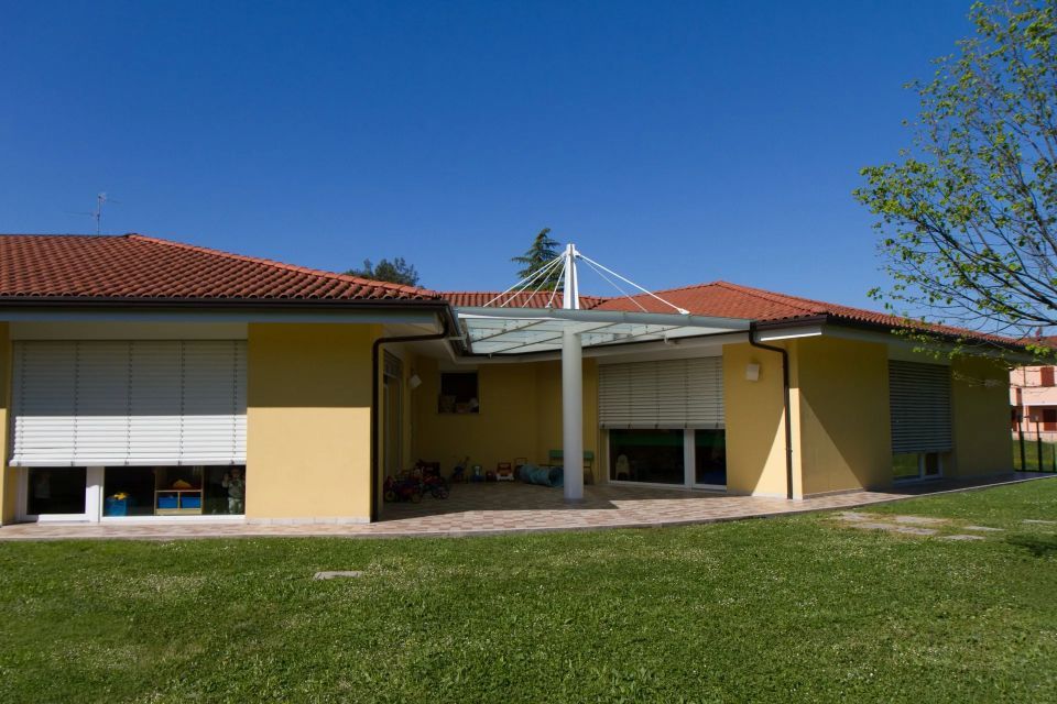 Edificio giallo con tetto di tegole arancioni, veranda e persiane chiuse su un prato verde sotto un cielo azzurro.