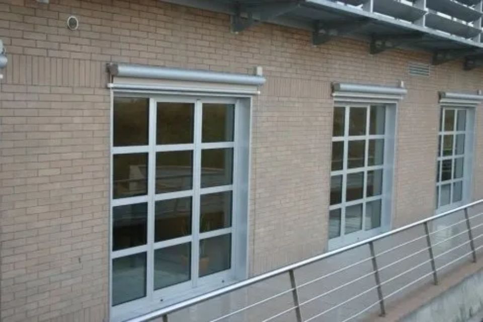 Facciata di un edificio in mattoni con tre finestre a griglia e tende avvolgibili argentate.