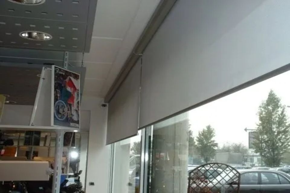 Vista della facciata interna del negozio con tende a rullo grigie parzialmente chiuse.