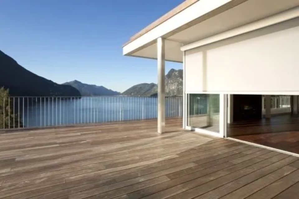 Terrazza in legno con vista sul lago e sulle montagne, con veranda coperta e tenda da sole bianca parzialmente abbassata.