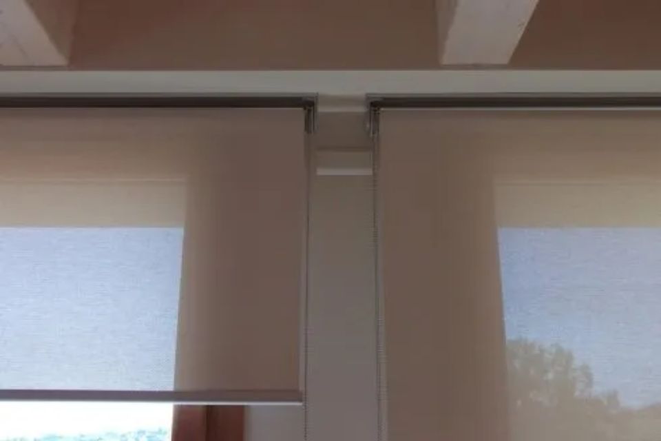 Due tende a rullo beige, parzialmente abbassate, montate su un binario bianco.