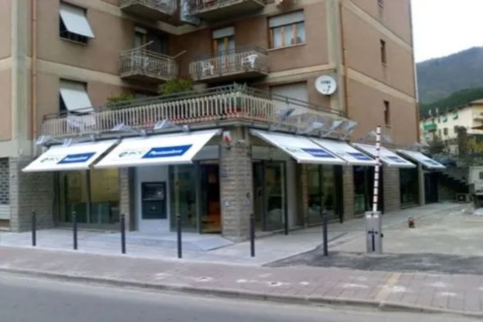 Filiale bancaria con tende da sole, vetrine e un palo di sbarramento su una strada cittadina.