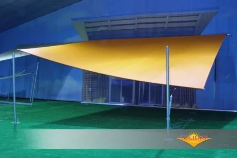 Vela ombreggiante triangolare gialla su superficie verde, fissata a pali e all'edificio.