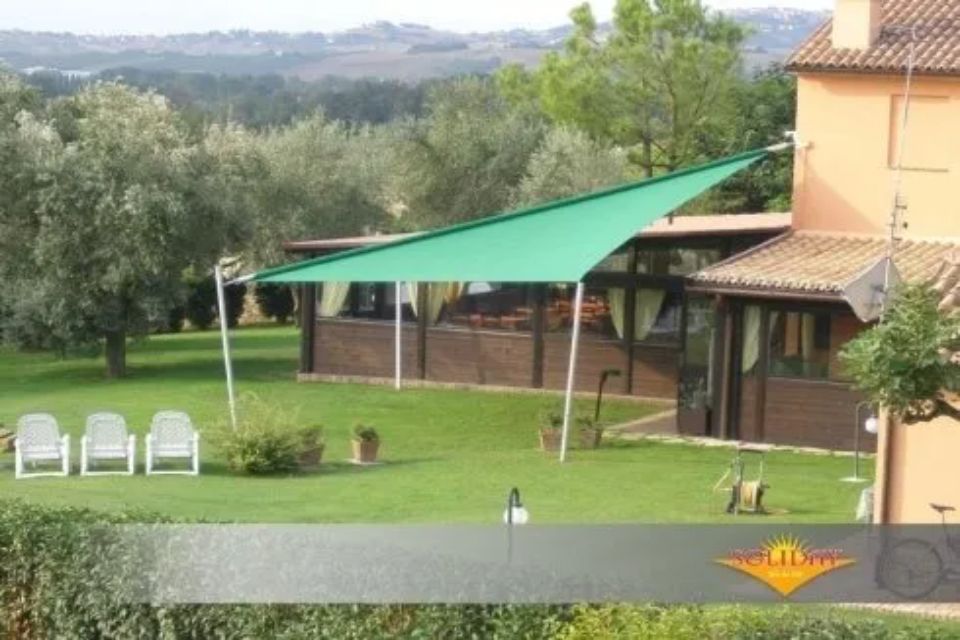 Tenda verde che copre un patio, un prato con sedie, alberi e un edificio sullo sfondo.