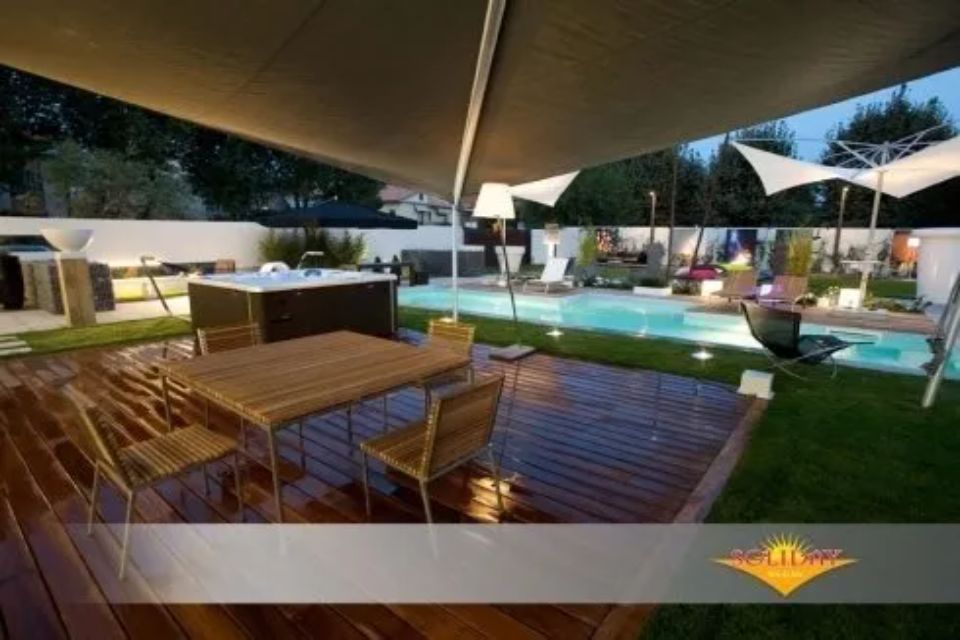 Patio esterno con terrazza in legno, tavolo, sedie, vasca idromassaggio, piscina e vele ombreggianti.