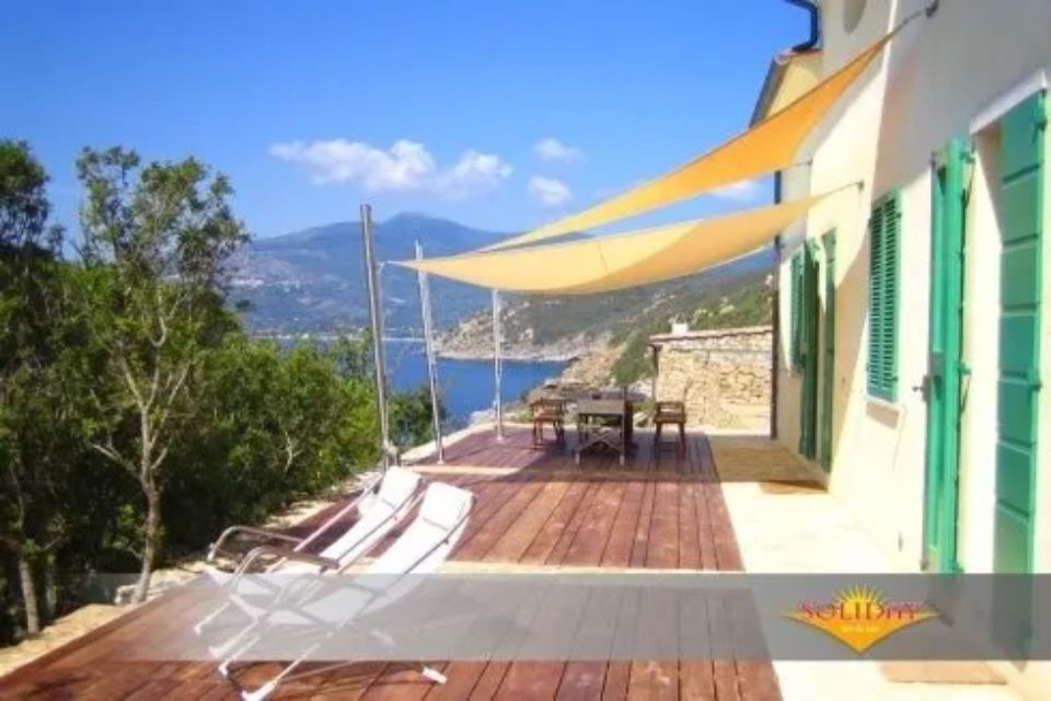 Patio con terrazza, tavolo e sedie con vista sul mare azzurro e sulle montagne, riparati da una vela ombreggiante.