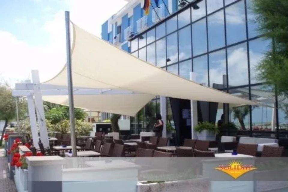 Un bar con tavoli e sedie sotto una vela bianca, di fronte a un edificio con grandi finestre.