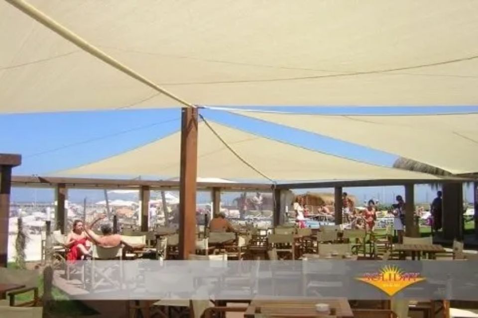 Ristorante all'aperto con vele ombreggianti color marrone chiaro, supporti in legno e clienti che cenano all'ombra.