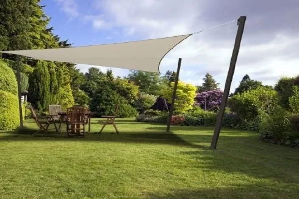 Una vela ombreggiante beige copre un tavolo e delle sedie in legno in un giardino erboso, sostenuta da pali scuri.