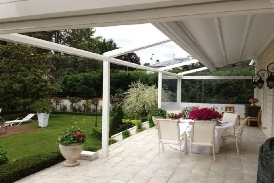 Patio bianco con tavolo, sedie, fiori e tenda da sole retrattile che si affaccia su un prato verde.