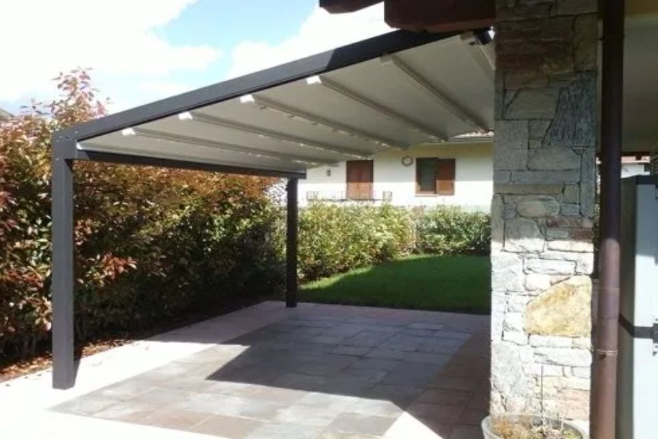 Pergola grigio scuro con tetto retrattile sopra un patio in pietra in un cortile con una casa visibile sullo sfondo.