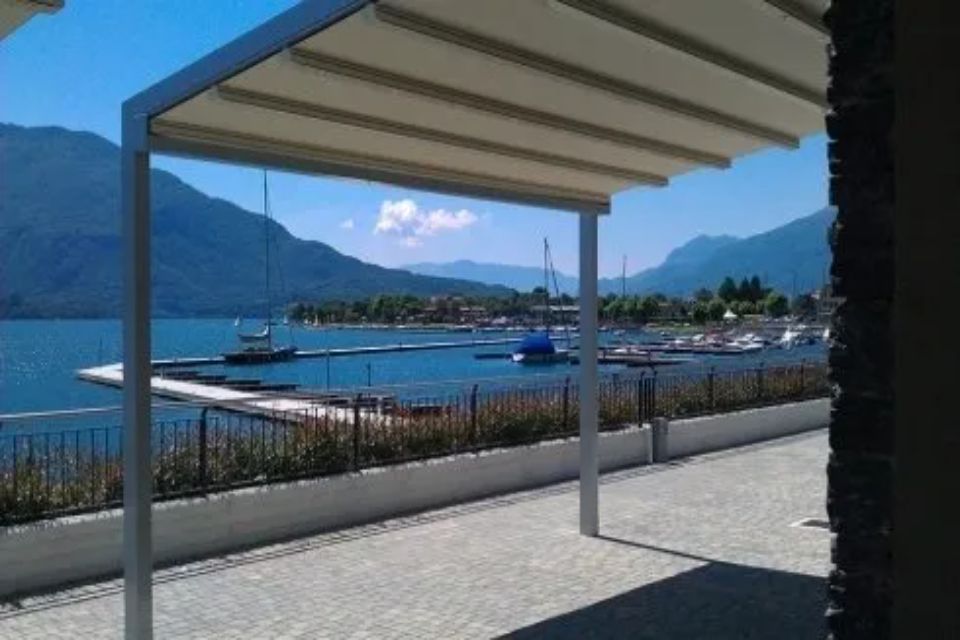 Pergola con vista su un lago con montagne, barche e una città sullo sfondo; giornata di sole.