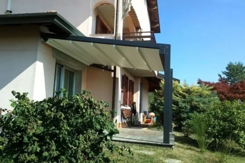 Pergola con tenda da sole retrattile fissata a una casa beige, giornata di sole.