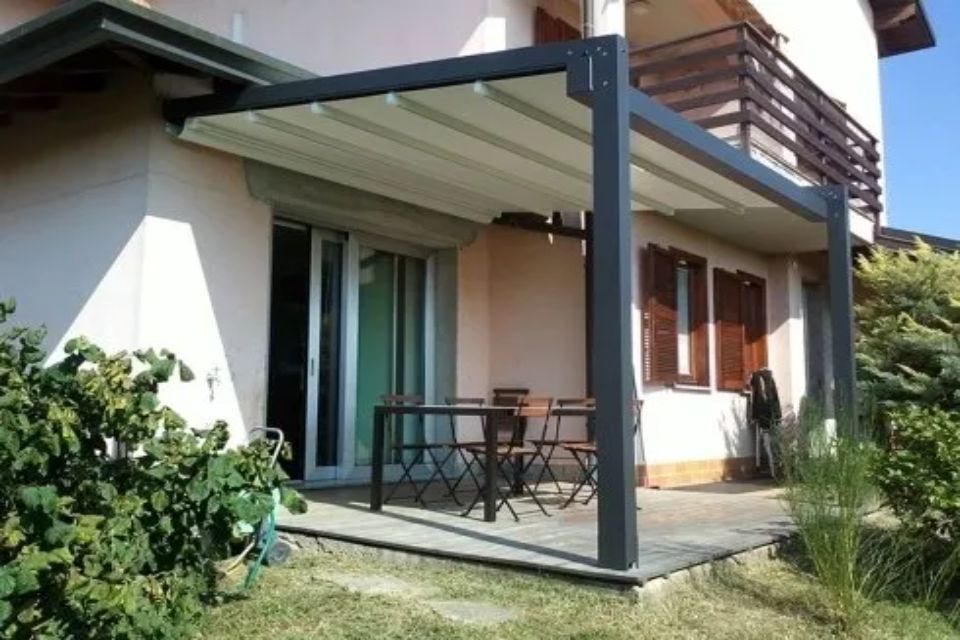 Patio con pergolato nero, tavolo, sedie e casetta beige con porta scorrevole in vetro.
