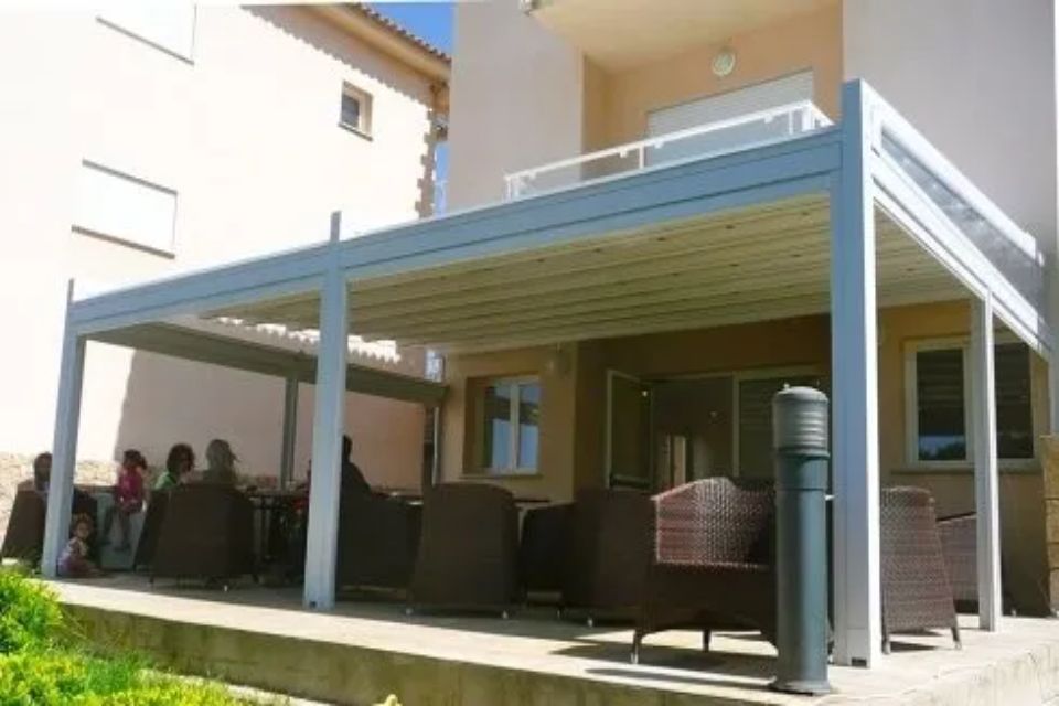 Patio con tetto retrattile e posti a sedere, persone riunite sotto. Edificio beige, soleggiato.